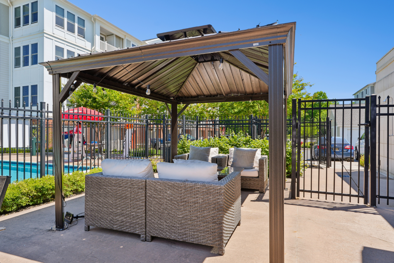 Poolside cabana with cushioned outdoor seating, shade structure, fencing, and apartment building behind it.