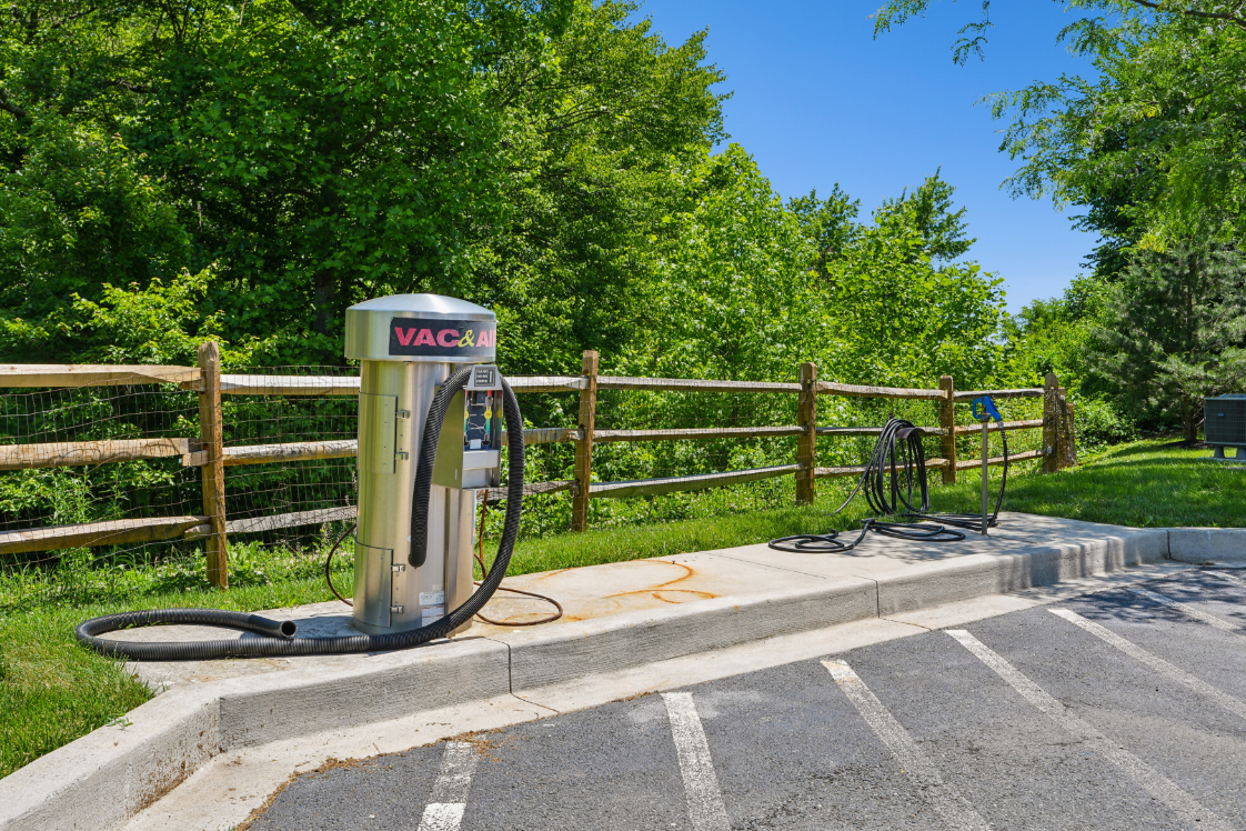 Outdoor car wash station with vacuum equipment, hose, parking spaces, fence, and trees.