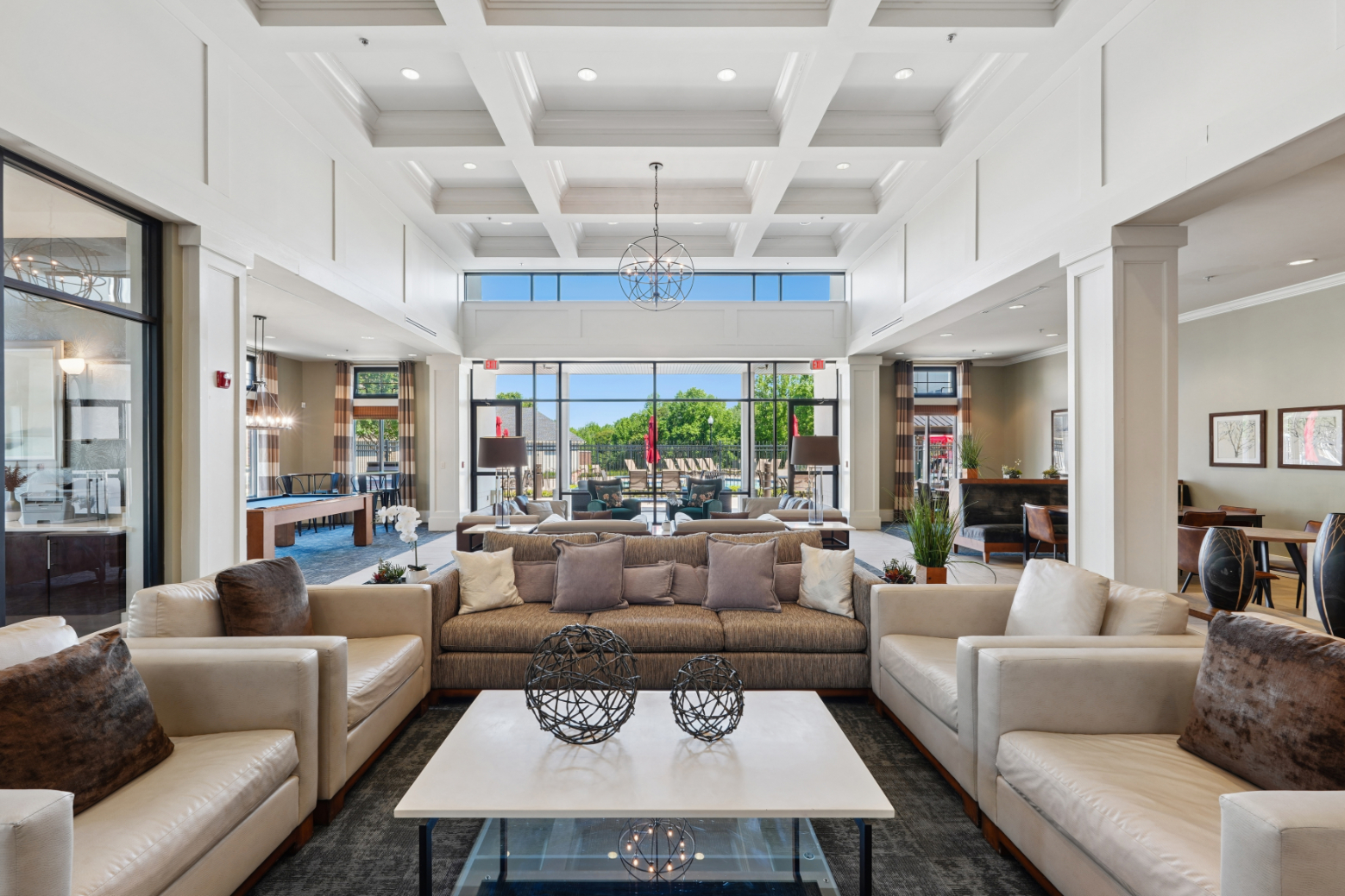 Bright clubroom with sofas, coffee tables, fireplace, large windows, and doors to the outdoor pool area.