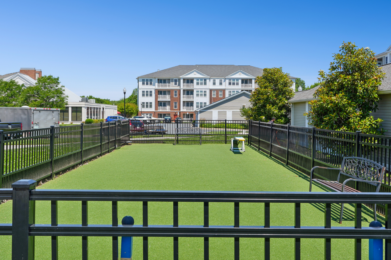 Fenced dog park with green turf, benches, dog agility features, trees, and apartment building in the background.