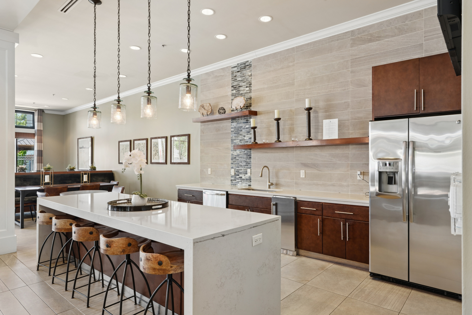 Entertainment kitchen with island seating, pendant lights, stainless refrigerator, wood cabinets, and tile backsplash.