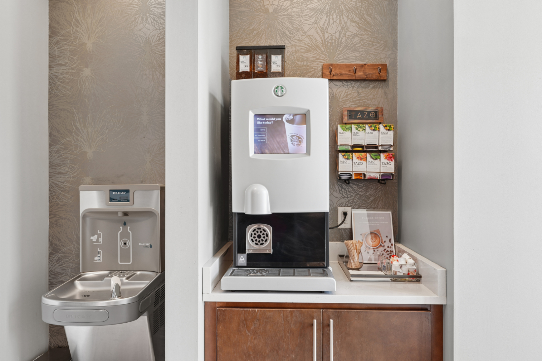 Hospitality station with coffee machine, water fountain, cups, snacks, and wood cabinetry.