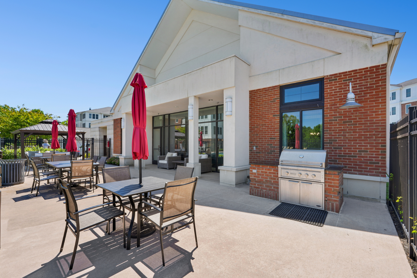 Outdoor grilling area with built-in grill, patio table, red umbrella, clubhouse, and pool fence.