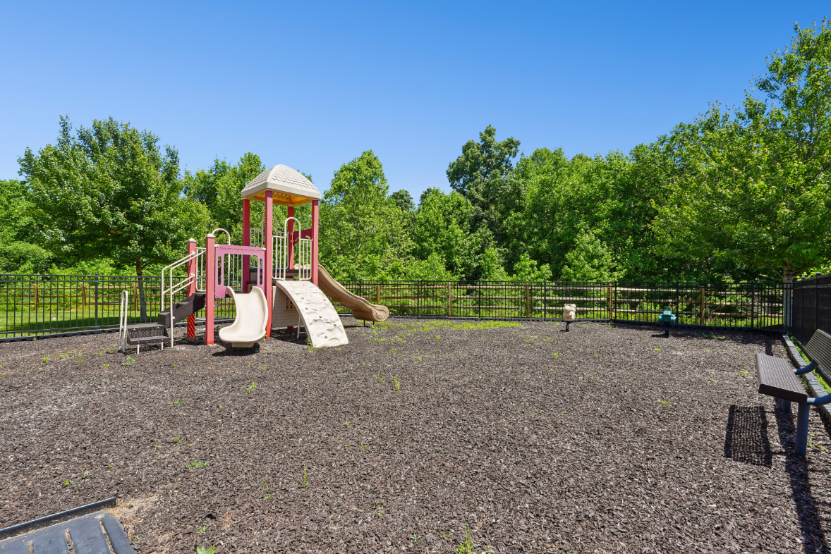 Outdoor playground with slides, climbing structure, mulch surface, benches, trees, and fencing.
