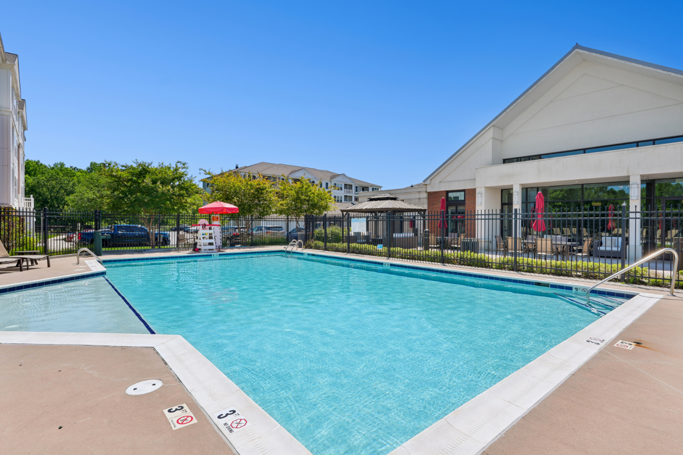 Outdoor swimming pool with lounge chairs, red umbrellas, fencing, and clubhouse building.