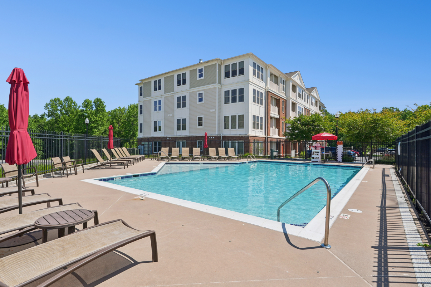 Outdoor pool area with lounge chairs, red umbrellas, apartment building, trees, and blue sky.