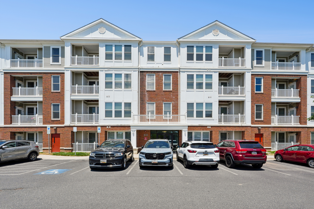 Exterior of a multi-story apartment building with balconies, brick accents, parked cars, and accessible parking spaces.