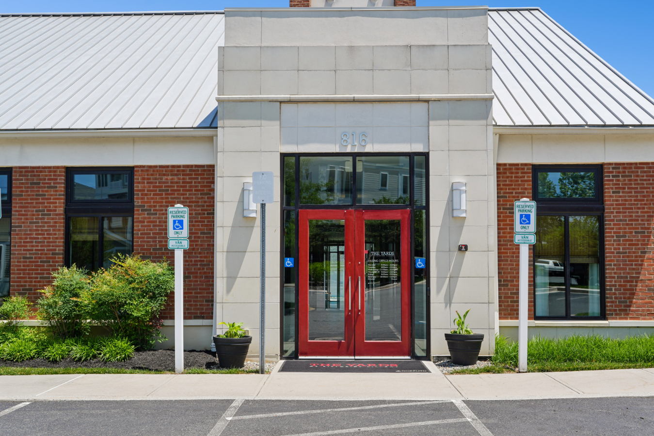 Leasing office entrance with red double doors, brick and stone facade, windows, planters, and accessible parking signs.
