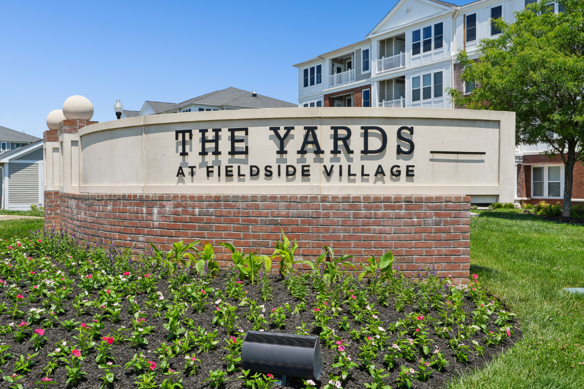 The Yards at Fieldside Village monument sign in front of flowers, brick base, lawn, and apartment building.