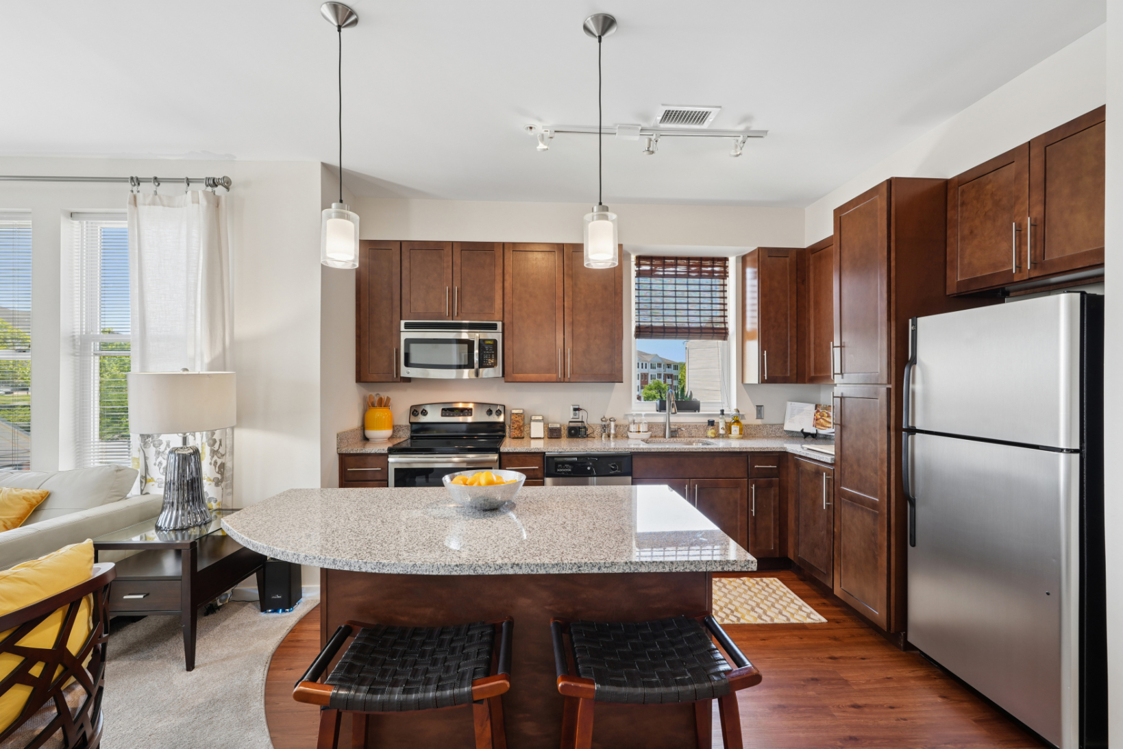 Apartment kitchen with stainless steel appliances, wood cabinets, granite countertops, pendant lights, and island seating.