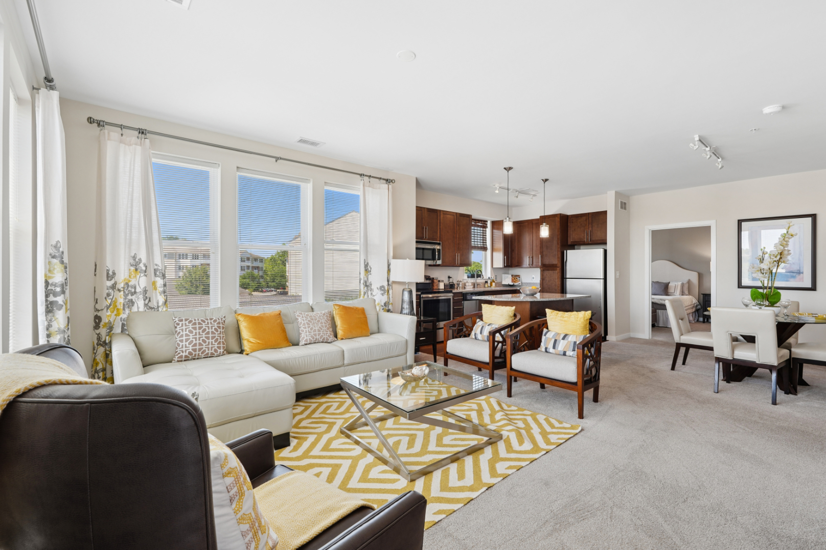 Spacious apartment living room with sofa, accent chairs, patterned rug, dining area, and kitchen in the background.