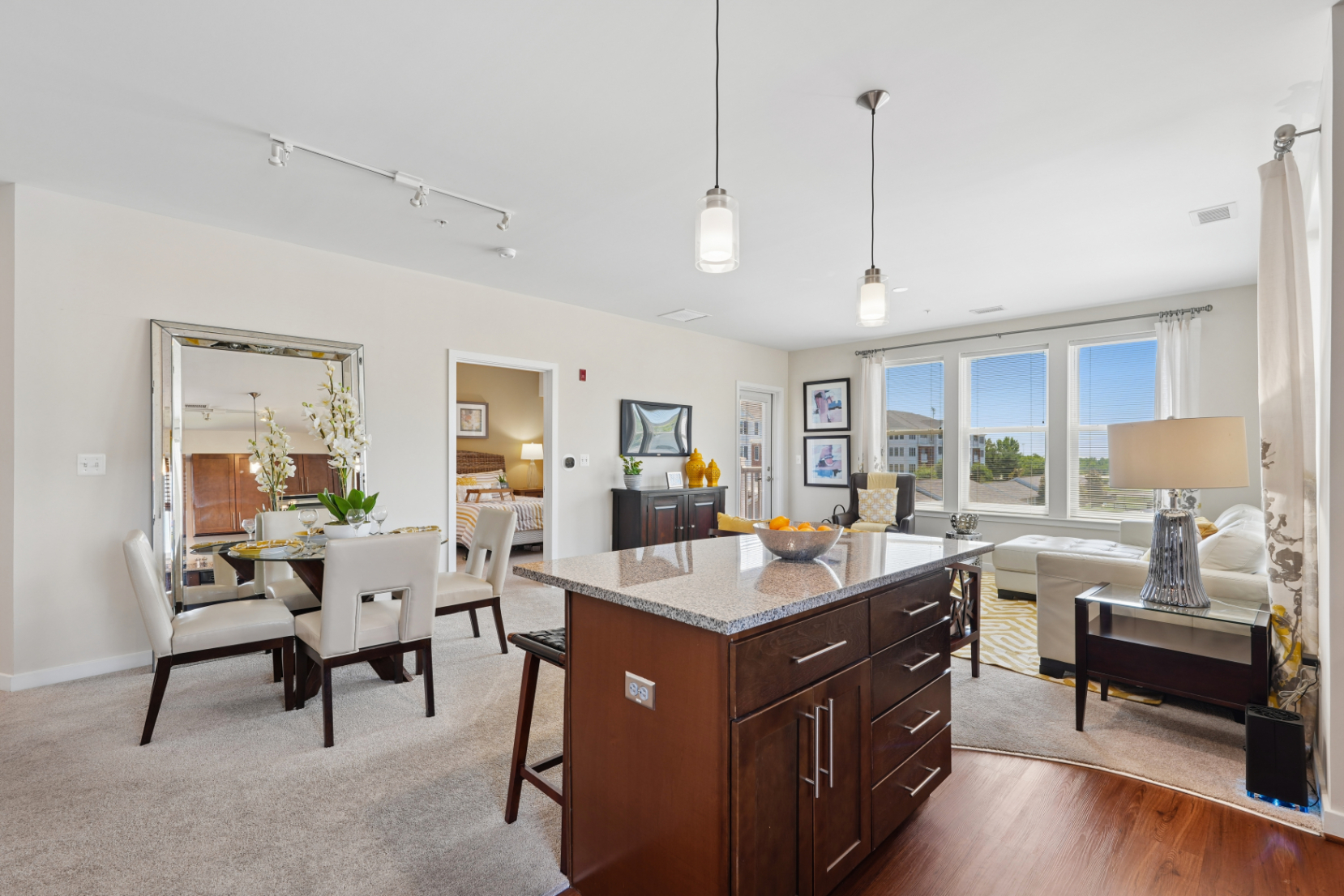 Open apartment layout with kitchen island, pendant lights, dining table, and living room seating near large windows.