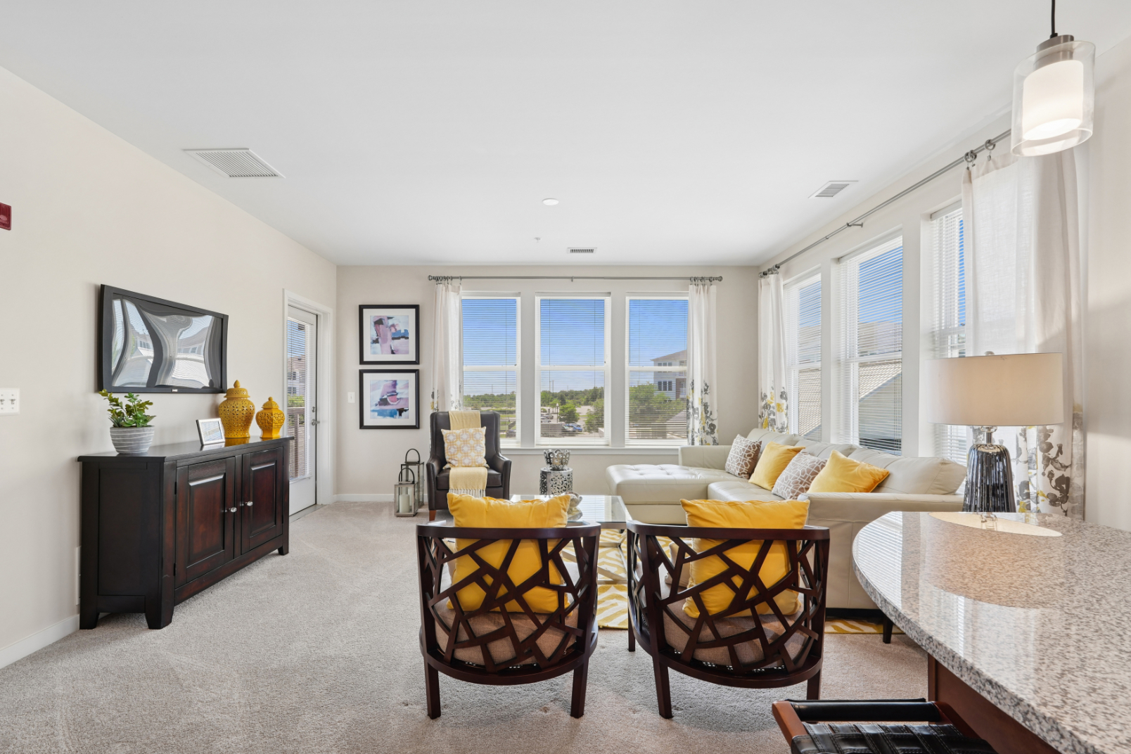 Bright apartment living room with sofa, accent chairs, dining table, large windows, and adjacent kitchen island.