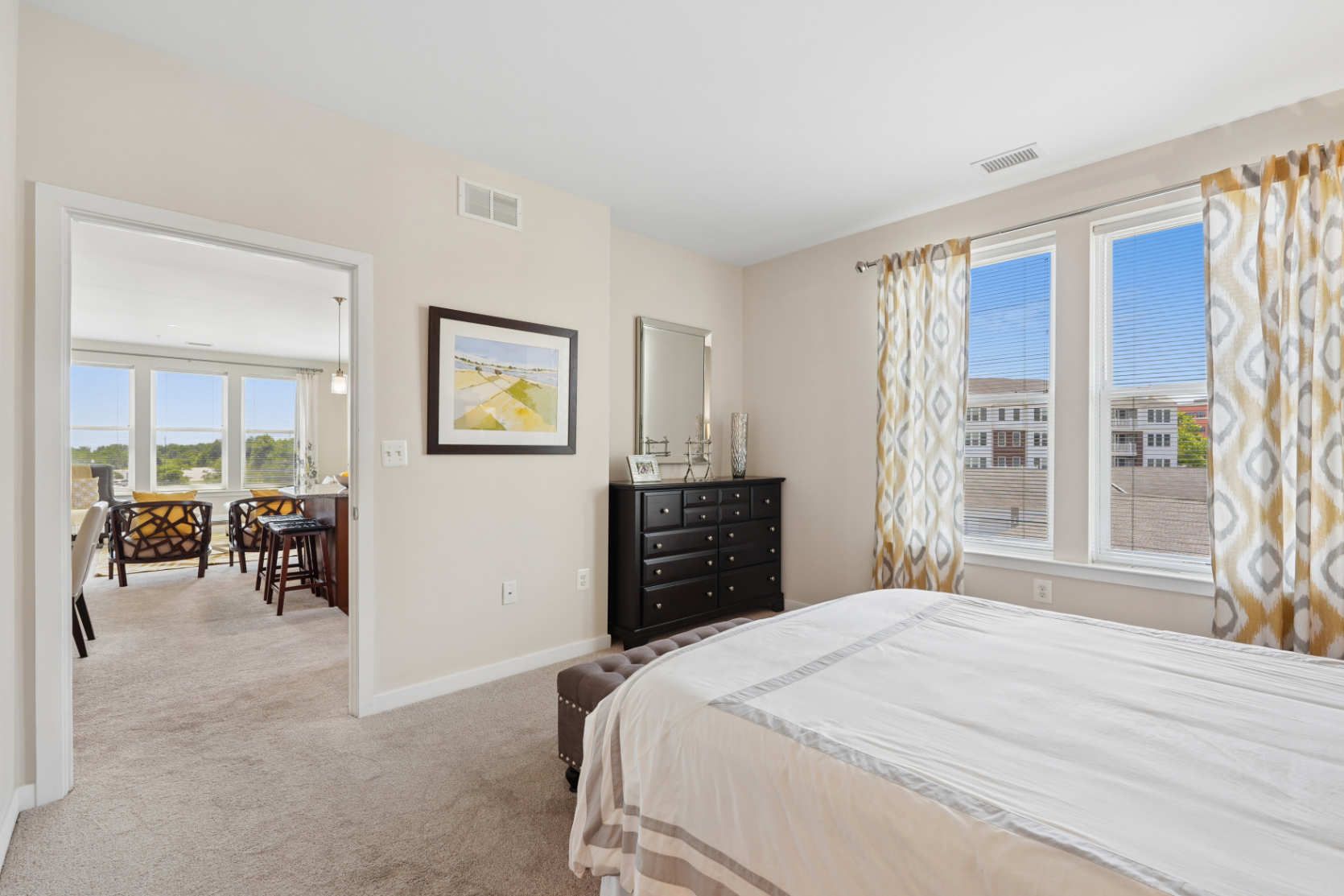 Bedroom with white bedding, dark dresser, yellow patterned curtains, and view toward the apartment living area.